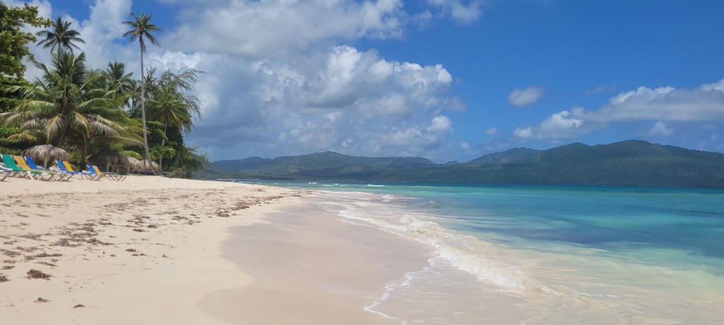 A serene beach scene on the Samaná peninsula, featuring golden sand, turquoise water, swaying palm trees, and a backdrop of lush mountains under a partly cloudy sky.