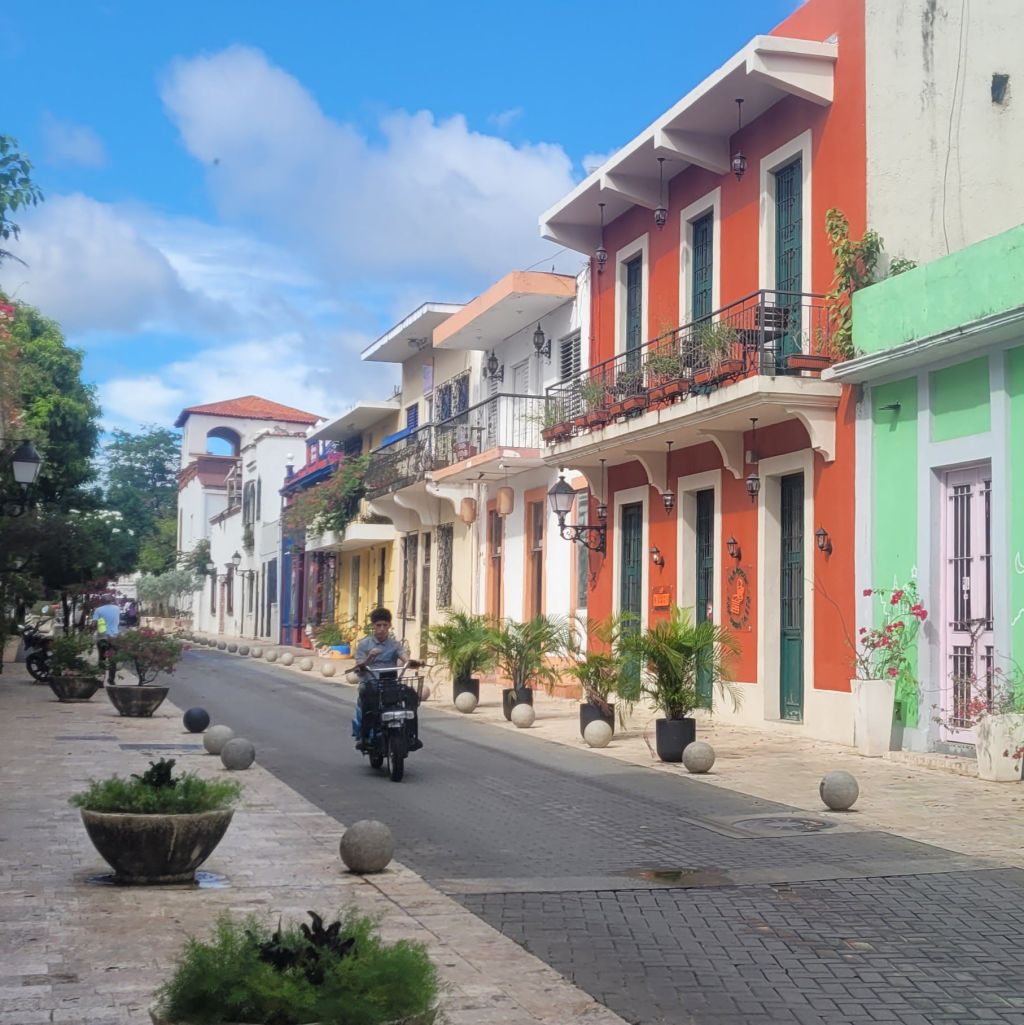 A charming street in the Zona Colonial, Santo Domingo, Dominican Republic featuring colorful colonial-style buildings, a clear blue sky, and a motorcyclist riding down the road.