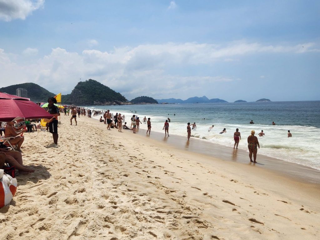 Crowded Copacabana beach in Rio de Janeiro, Brazil with people enjoying the sun, some lounging under umbrellas, while others are playing in the waves. Hills and distant islands are visible in the background under a partly cloudy sky.