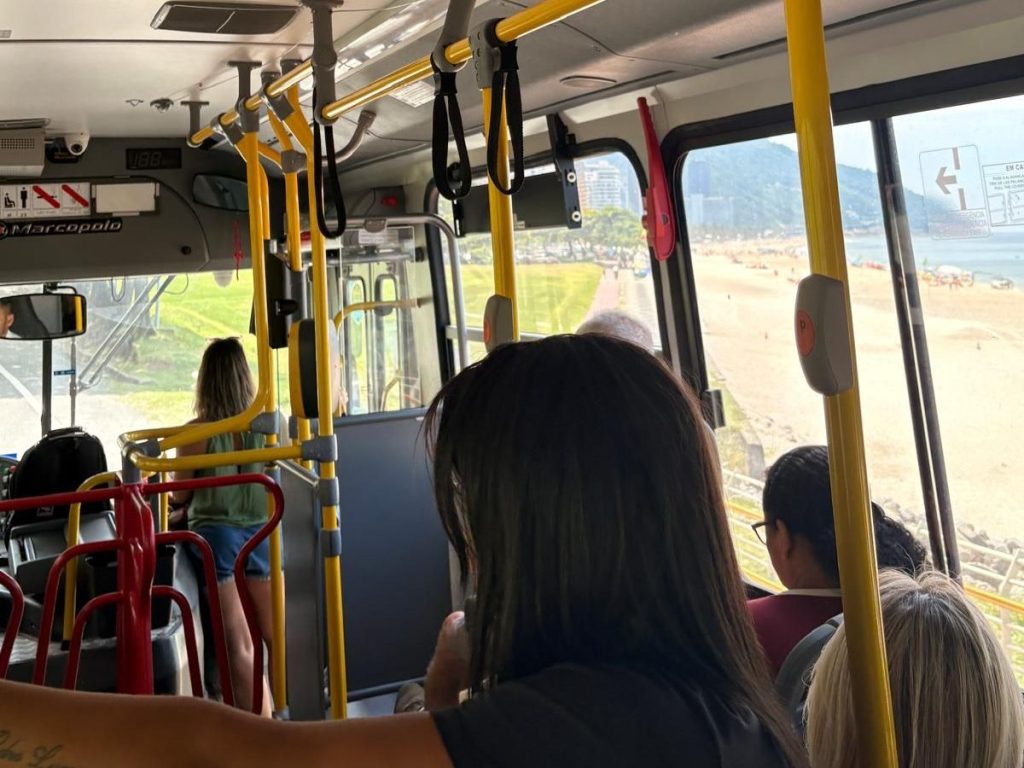Interior view of a bus with passengers, showing yellow handrails and a beach outside through the windows.