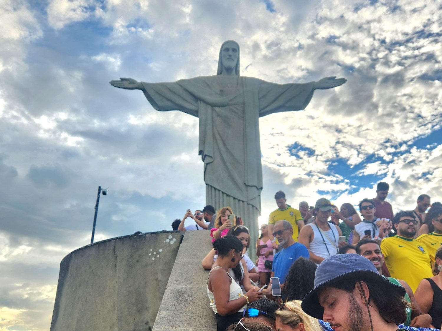 Crowd of people gathered around the Christ the Redeemer statue against a cloudy sky.