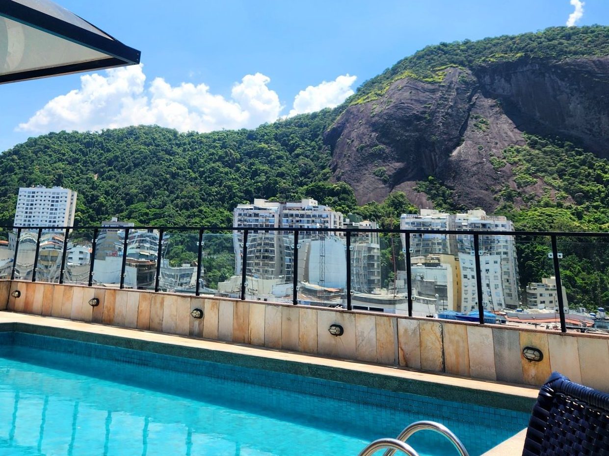 A scenic view from Copacabana Mar Rooftop pool overlooking lush green mountains and city buildings under a blue sky with fluffy white clouds. Taken in Rio de Janeiro, Brazil.