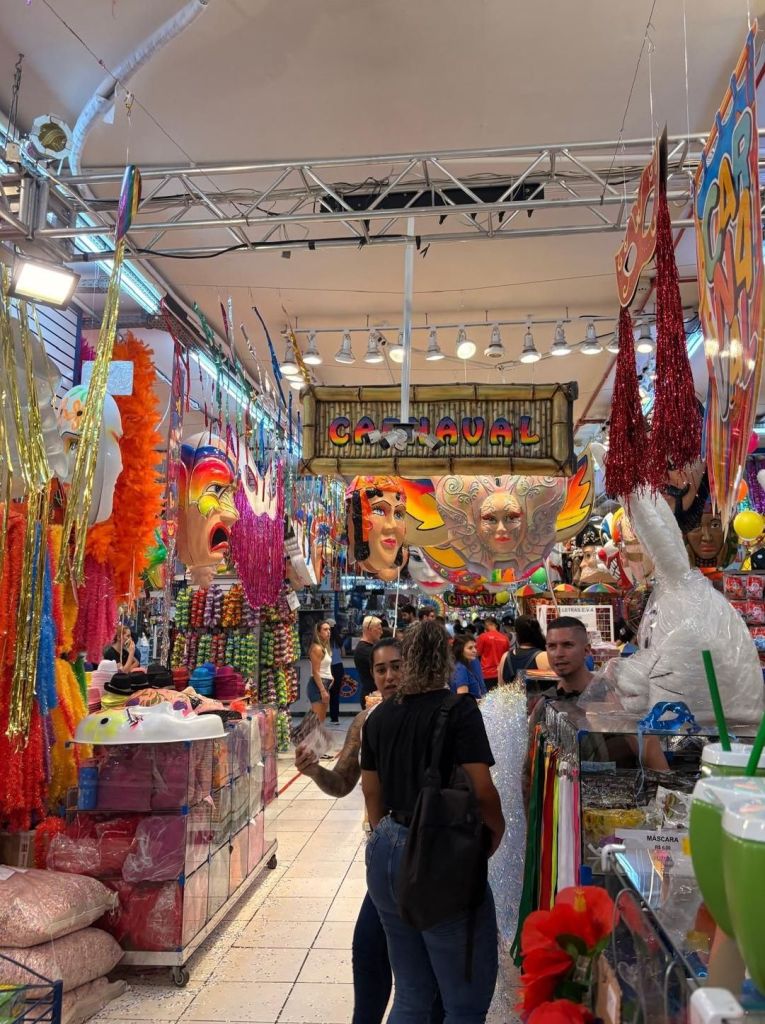 A vibrant market in Rio filled with colorful decorations, costumes, and masks for a carnival theme, featuring several people browsing and interacting.