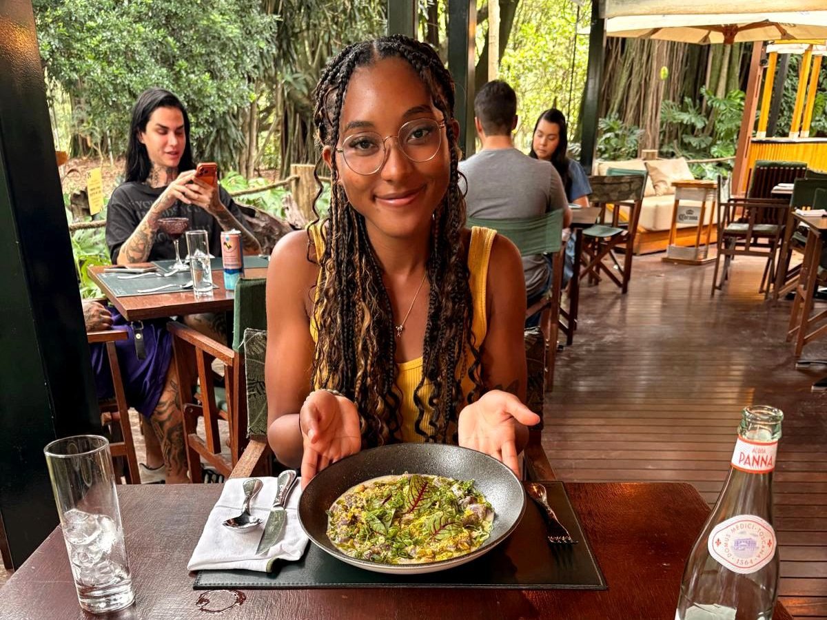 A smiling woman with braided hair sits at a restaurant table, presenting a plate of food. In the background, other diners are seated, enjoying their meals in a lush, green setting.