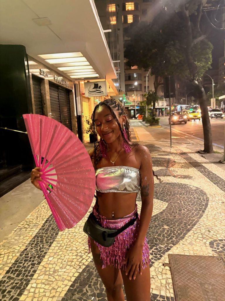 A woman with braided hair stands on a city street in Copacabana, Rio at night, holding a pink fan. She wears a silver crop top and a fringed mini skirt, accessorized with a small black fanny pack.