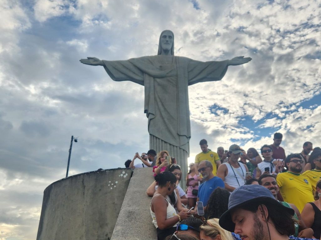 Crowd of people gathered around the Christ the Redeemer statue against a cloudy sky.