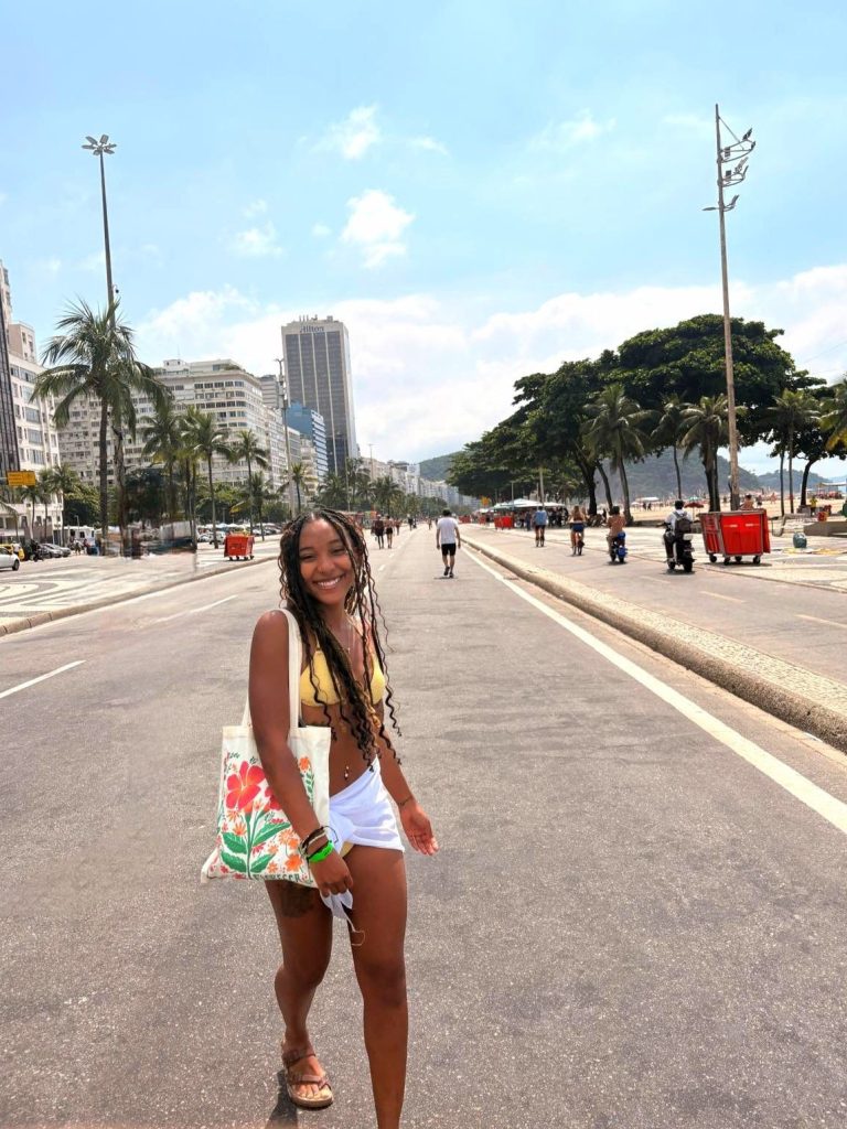 A young woman with long, braided hair wearing a yellow bikini top and white cover-up smiles while walking along Copacabana boardwalk in Rio de Janeiro, Brazil. She holds a colorful tote bag, with palm trees and city buildings in the background under a clear blue sky.