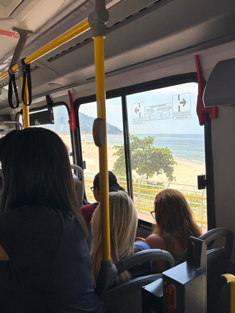View from inside a bus in Rio showing passengers looking out the window at a beach and ocean landscape.
